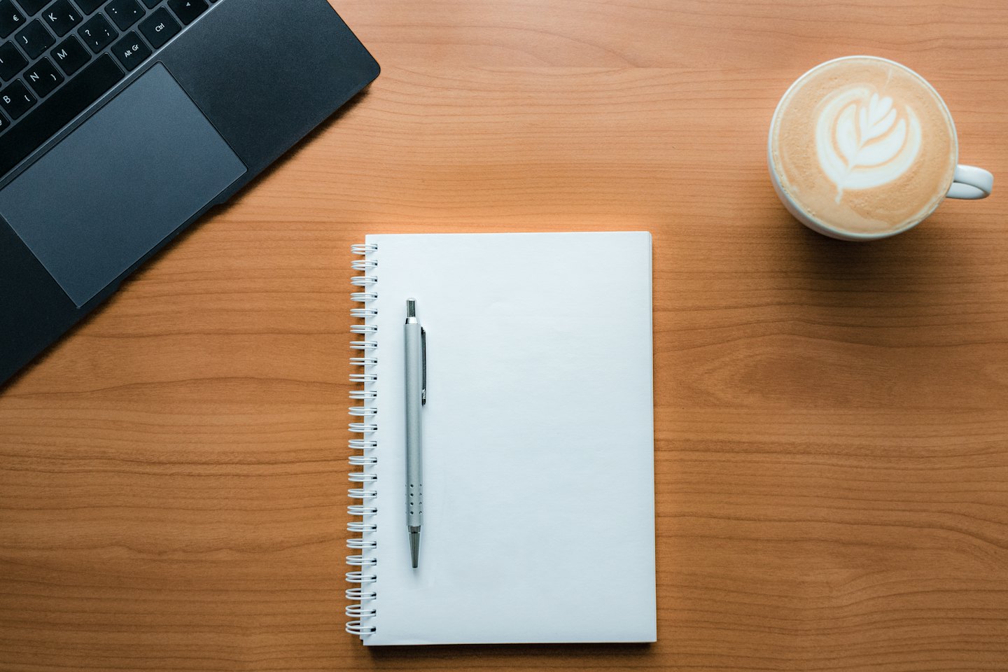 Laptop, notebook, pen, and coffee cup on a wooden desk.